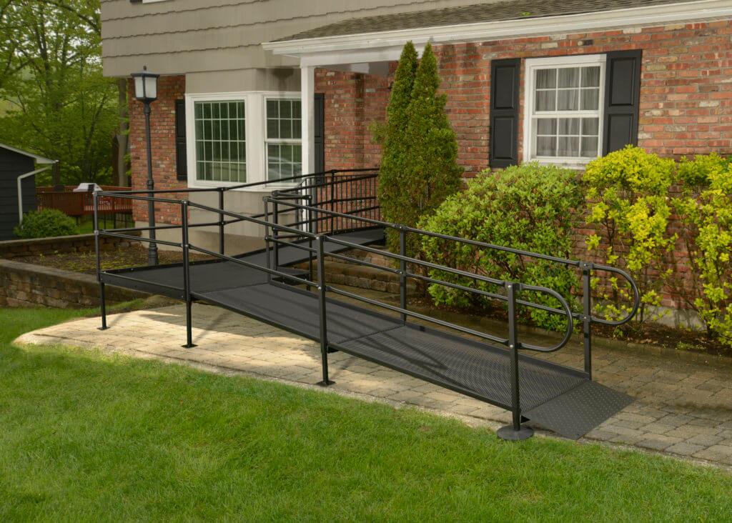 A sleek black metal accessibility ramp, known for its cost-effectiveness, leads to the front entrance of a charming brick house with white windows and black shutters, all embraced by lush greenery.