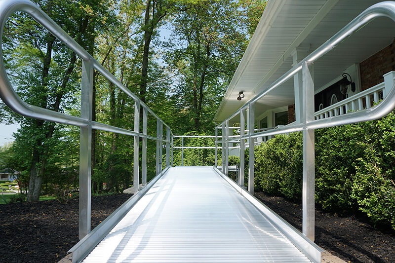 A metal accessibility ramp leads to the entrance of a house surrounded by greenery, seamlessly complementing the stairlifts inside.