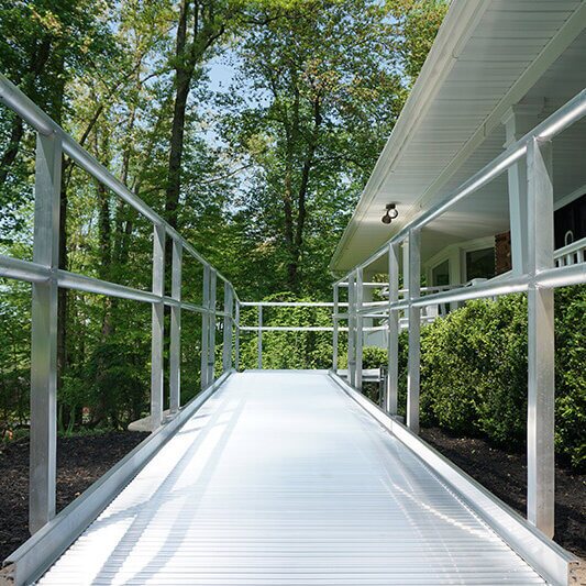 A metal accessibility ramp leads to the entrance of a house surrounded by greenery, seamlessly complementing the stairlifts inside.