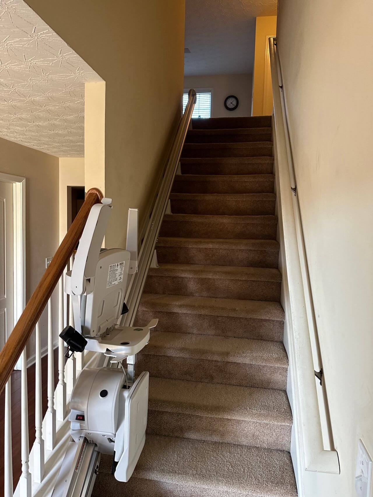 A stair lift is installed on a residential staircase, with beige carpeted steps and a light yellow wall. The lift includes a rail, seat, and mechanical controls. Natural light comes from a window above.
