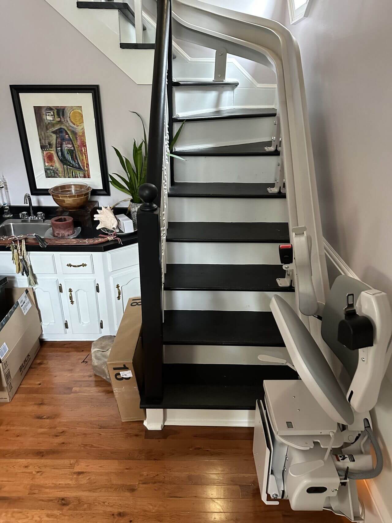 A staircase with a stairlift installed on the side. Boxes are placed under the stairs, and a cabinet with decor is on the left side.