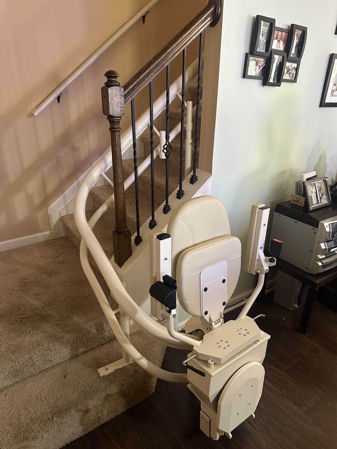 A stairlift with a beige seat is installed along a curved staircase with wooden railings, next to a wall displaying framed photos.