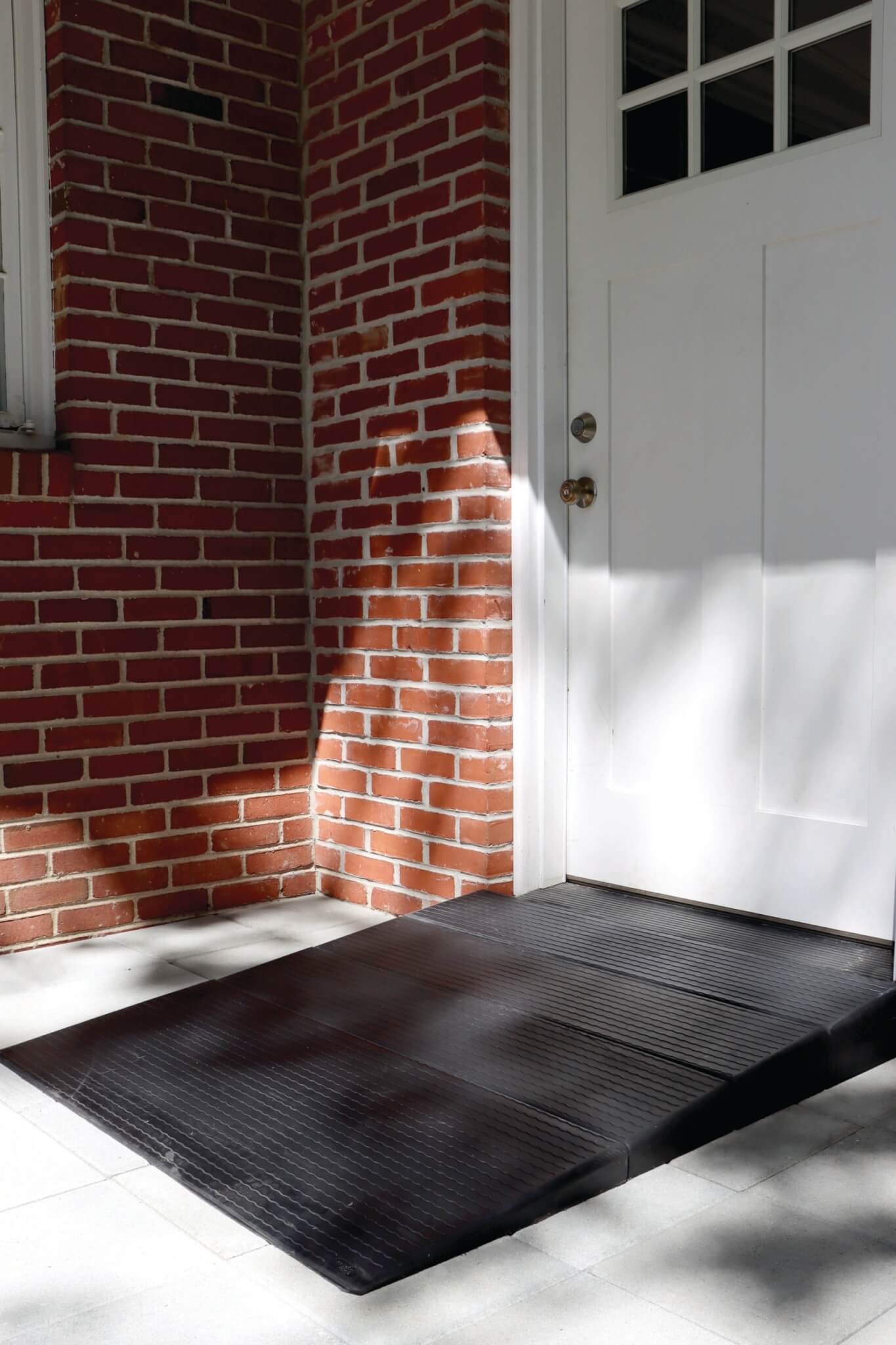 A rubber ramp leads up to a white door set in a red brick wall, with sunlight casting shadows on the tiled ground.