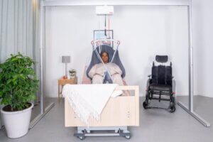 A person lies in a hospital bed using a patient lift system, with a wheelchair nearby and a potted plant and nightstand beside the bed.