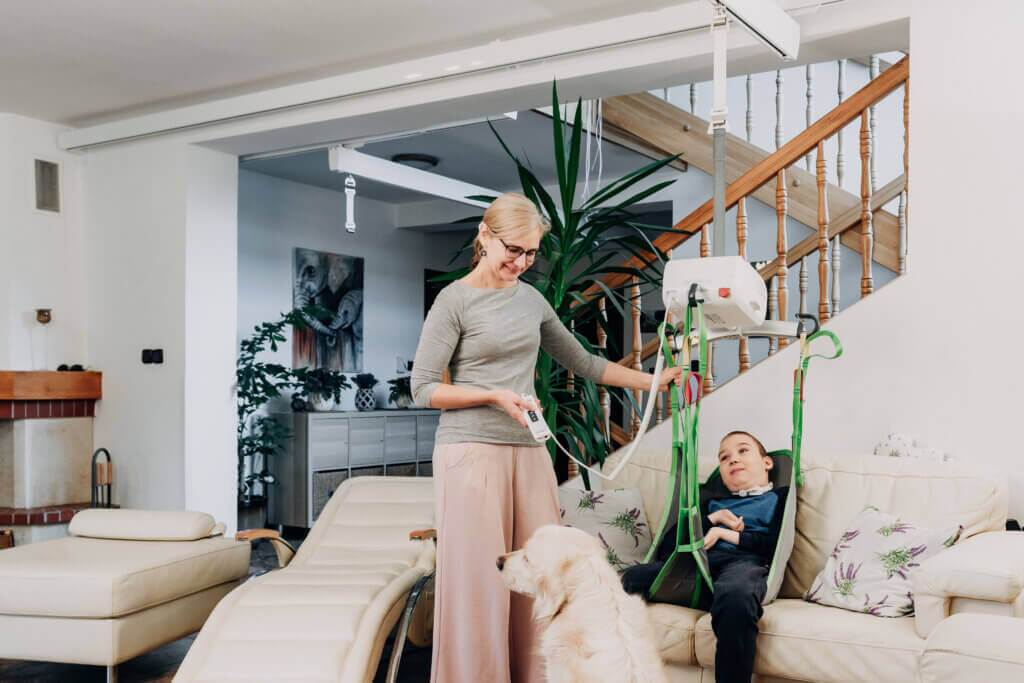 A woman operates a ceiling lift to assist a child in a harness chair in a living room, with a dog nearby.