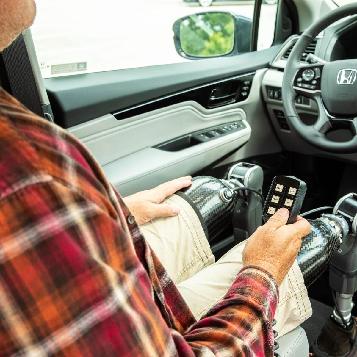 Honda_6way_UWR_2019_010_High A person with prosthetic legs sits in the drivers seat of a car, holding a remote control device, with the car door open.