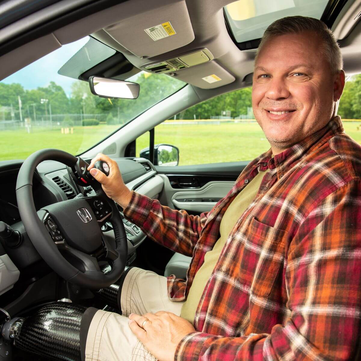 Honda_6way_UWR_2019_013_High (1) A man with a prosthetic leg sits in the driver’s seat of a car, smiling, with his hand on the steering wheel. A park is visible through the window.