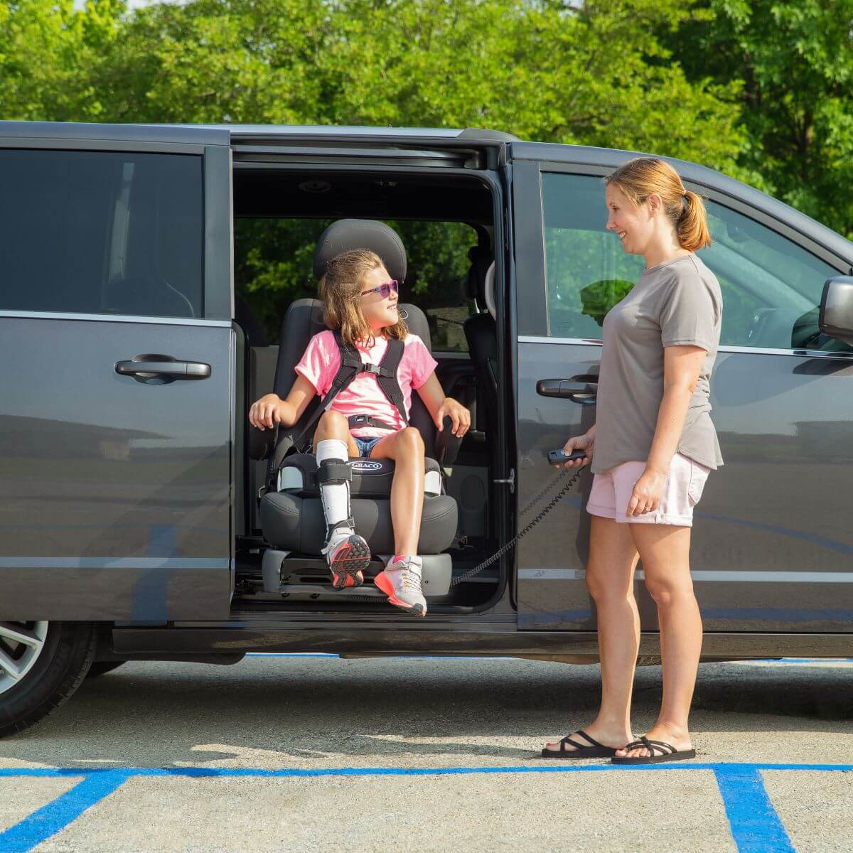 Turny_2019_Dodge_Mid_002_High A woman uses a remote control to operate a lift seat, helping a young girl with a leg brace exit a minivan in a parking lot.