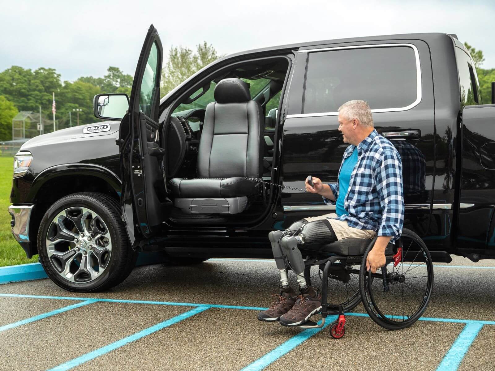 Turny_2019_Truck_011_High5 A man in a wheelchair with prosthetic legs uses a remote to operate a modified truck featuring transfer seats and a rotating driver's seat in an accessible parking space.