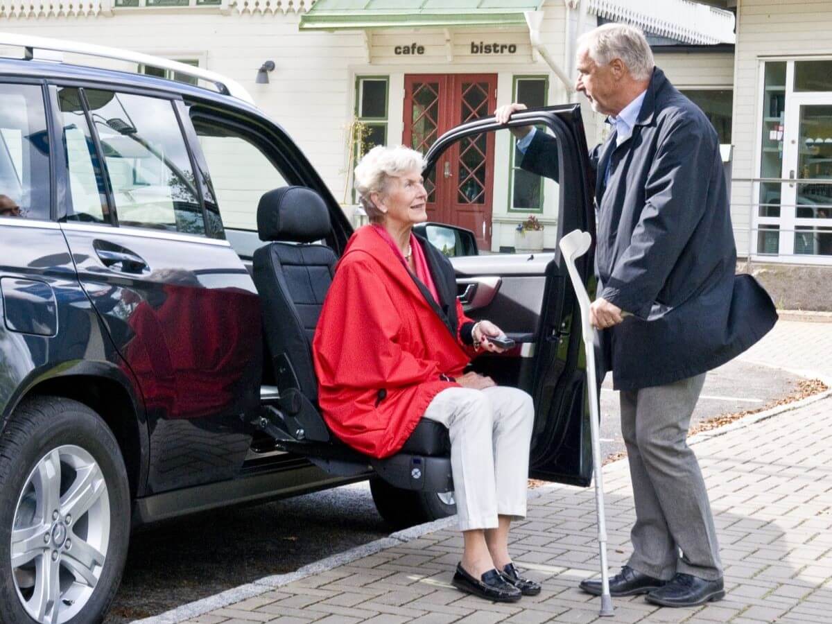 Turny_Evo_Lifestyle_High_1 An older woman sits on a swivel car seat while an older man with a cane stands beside her in a parking lot outside a cafe and bistro.