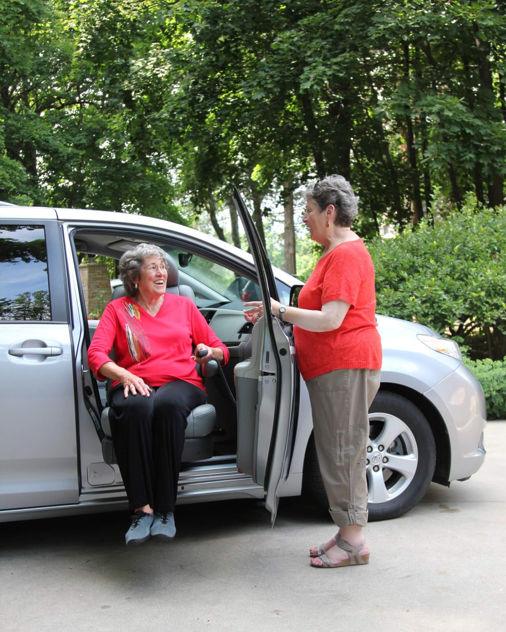 Turny_Evo_Woman_Caretaker_High An older woman sits on a cars swivel seat while another woman stands by the open car door, both smiling, in a driveway surrounded by greenery.