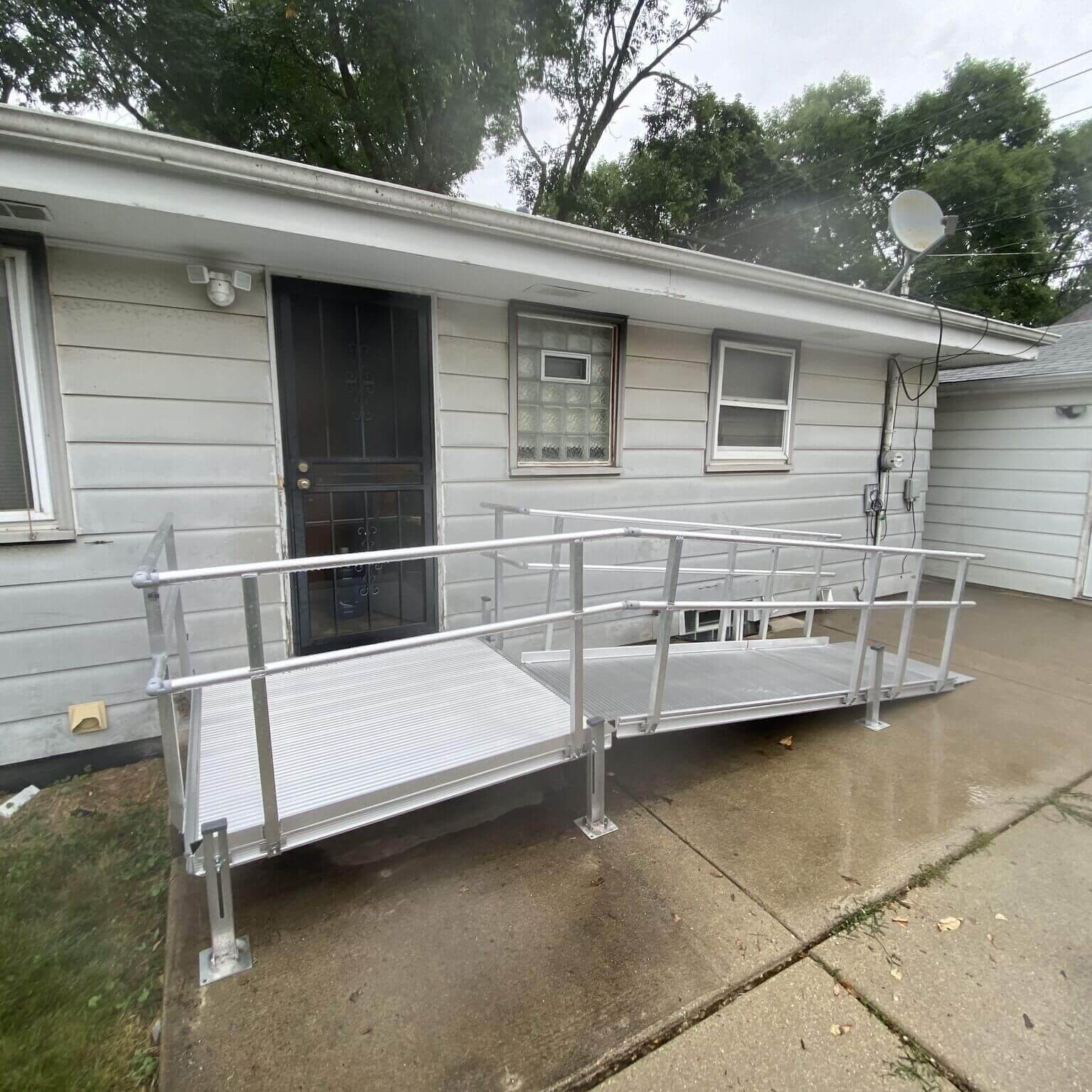 An aluminum wheelchair ramp leads up to a gray houses back door, set on a concrete patio. Trees and additional buildings are visible in the background.