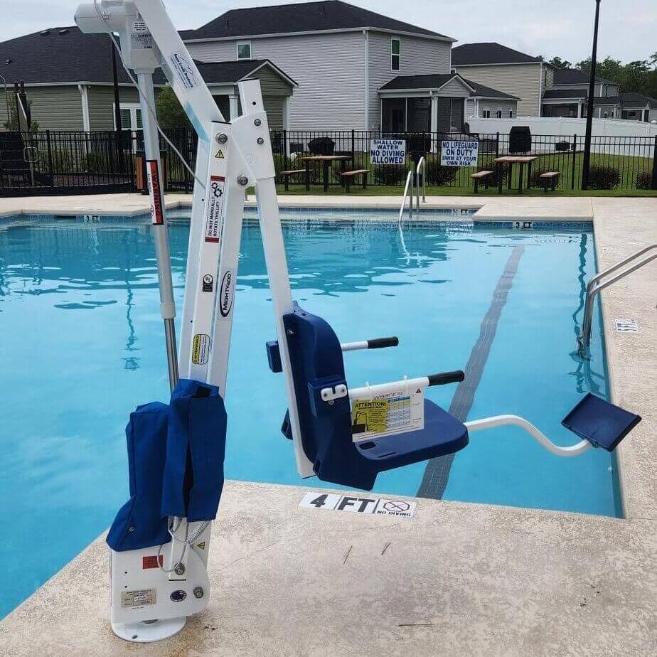A pool lift chair by the edge of a swimming pool, designed for accessibility. Houses are visible in the background.