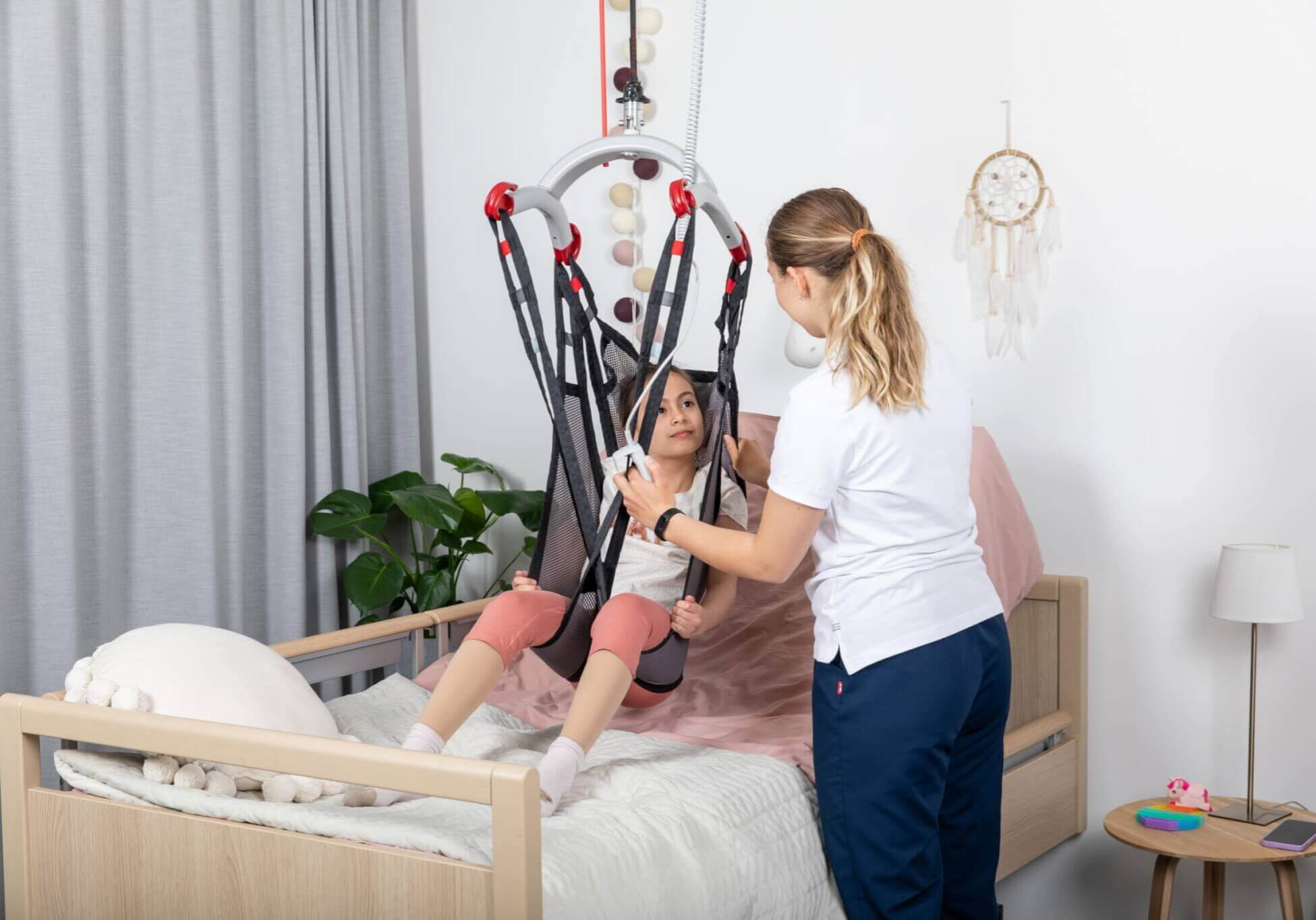 A caregiver assists a young girl in a lift sling above a bed in a well-lit room with a dreamcatcher and plant.