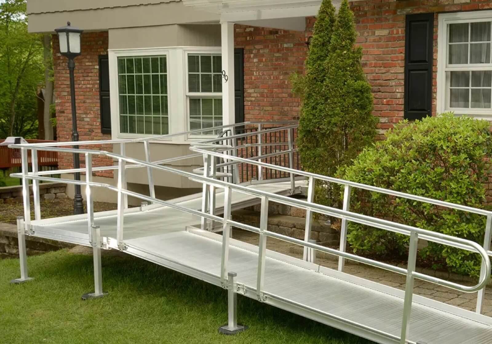 The aluminum wheelchair ramp for home leads gracefully to the entrance of a brick house, complemented by charming white windows and lush green shrubs.