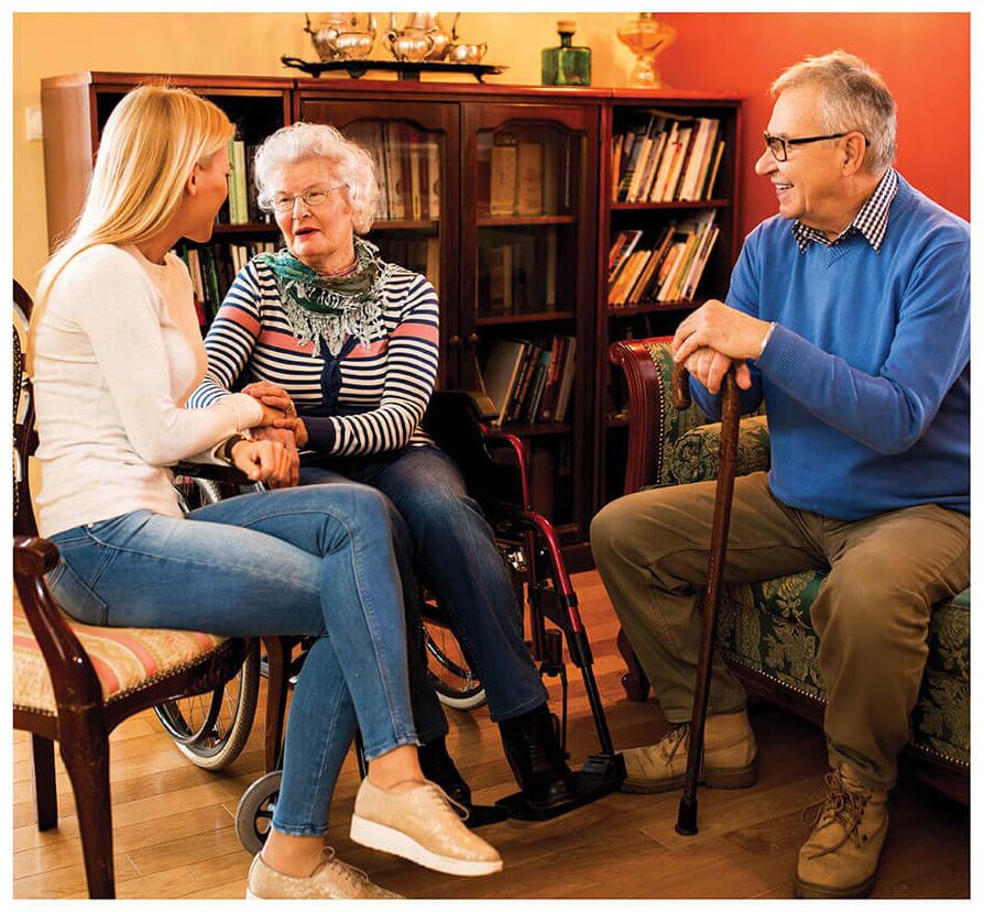 A young woman holds hands with an elderly woman in a wheelchair from MobilityWorks, while an older man with a cane sits nearby in a cozy room filled with shelves and books.
