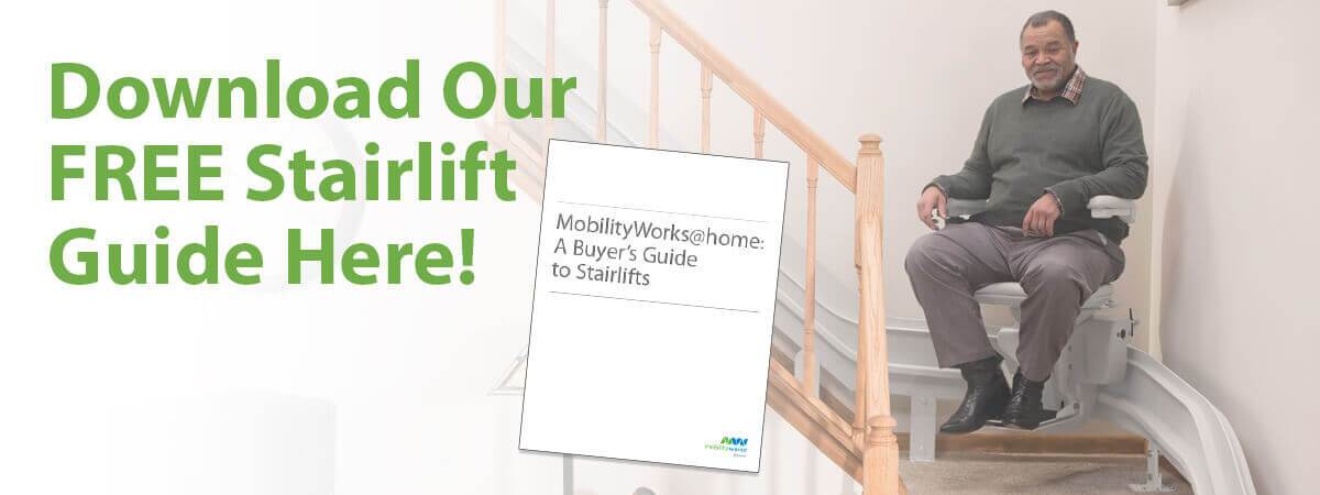 A man sits on a stairlift near a staircase with a sign reading Download Our FREE Stairlift Guide Here! and a guide titled Mobilityworks@home: A Buyers Guide to Stairlifts.