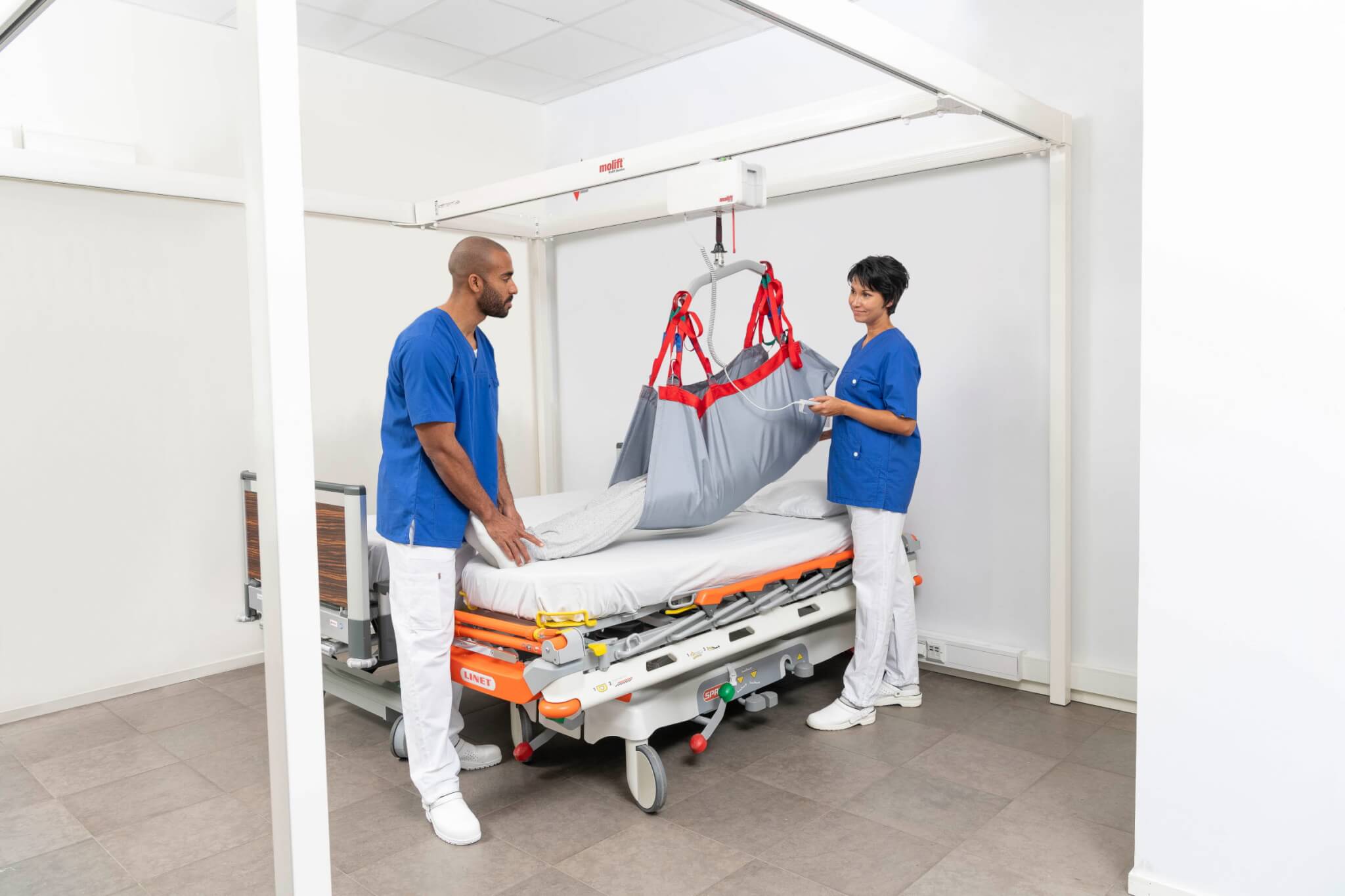 Two healthcare workers use a mechanical lift to move a patient from a hospital bed. They wear blue scrubs and are in a medical facility.