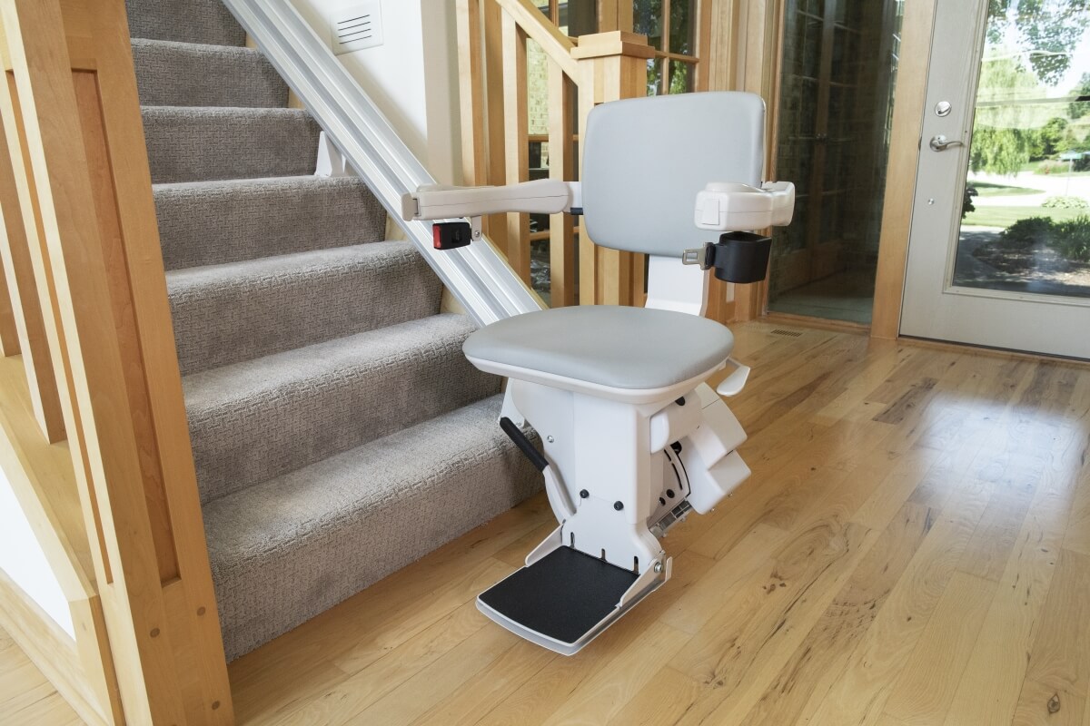 A stairlift is installed alongside a residential staircase, featuring a gray seat and armrests.