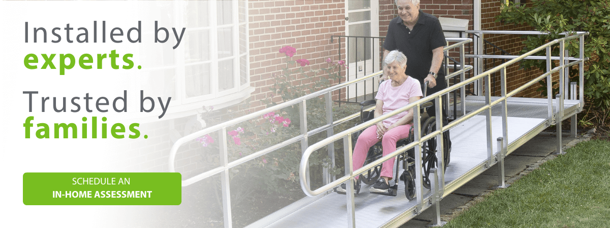 An older man pushes a woman in a wheelchair down an outdoor metal ramp beside a brick house. Text reads, Installed by experts. Trusted by families. Schedule an in-home assessment.