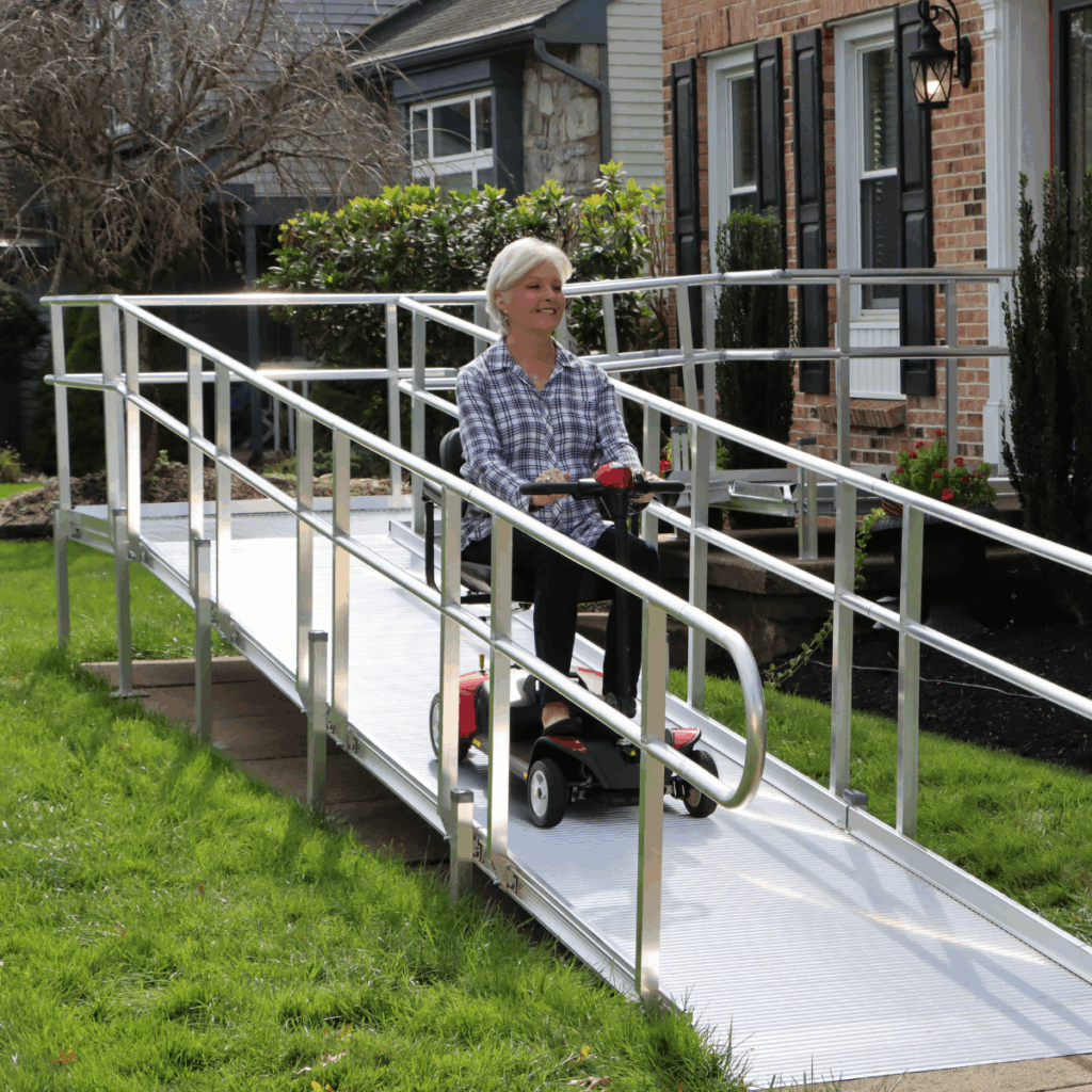 An older woman rides a mobility scooter down a metal wheelchair ramp outside a brick house on a sunny day.