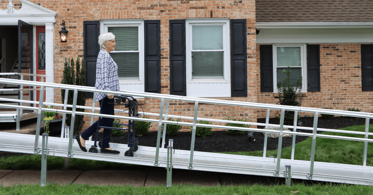 An older adult using a walker moves up a metal wheelchair ramp outside a brick house with black shutters.