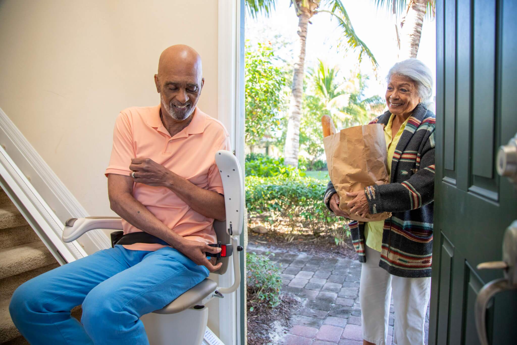 An older man sits on a stair lift by the stairs inside a house, while an older woman enters through the door holding a paper bag of groceries.