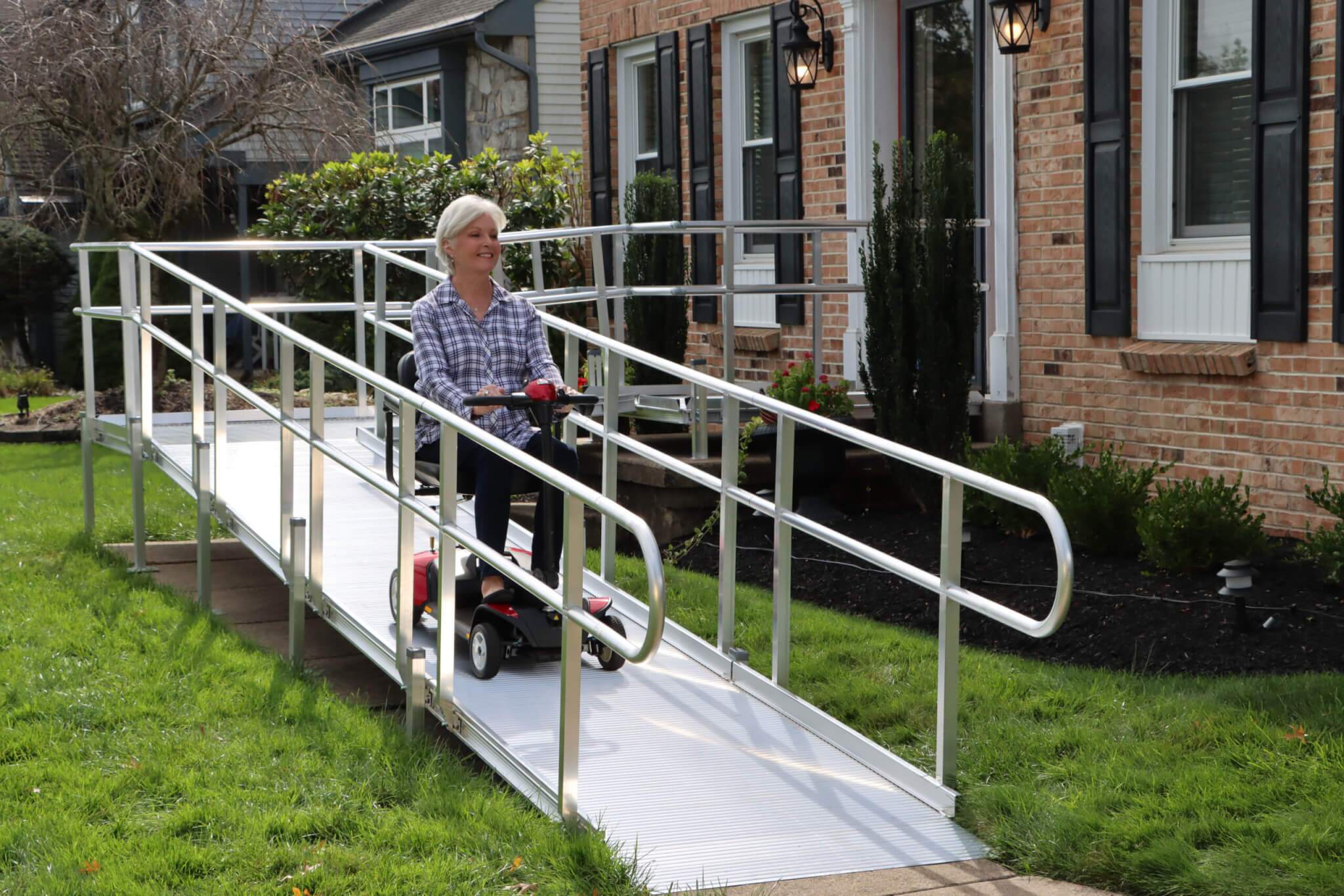 An older woman rides a red mobility scooter down a metal ramp attached to the front entrance of a brick house.
