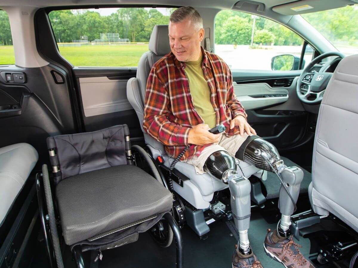 A man with two prosthetic legs sits in the drivers seat of a car beside his folded wheelchair, holding a remote control and looking at the wheelchair.
