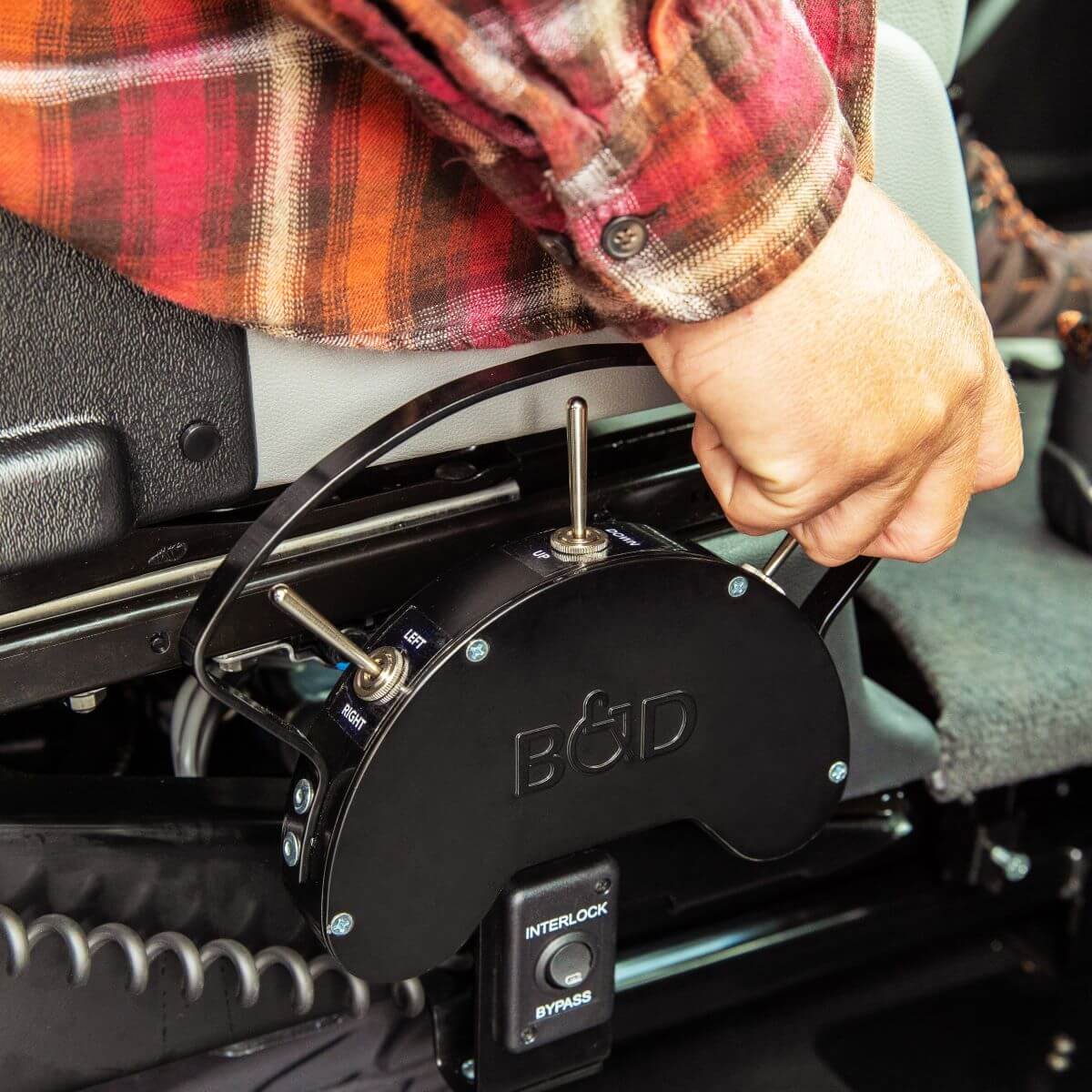 A person operates a B&D-branded mechanical control lever mounted beside a vehicle seat, with switches labeled “Interlock” and “Bypass” visible.