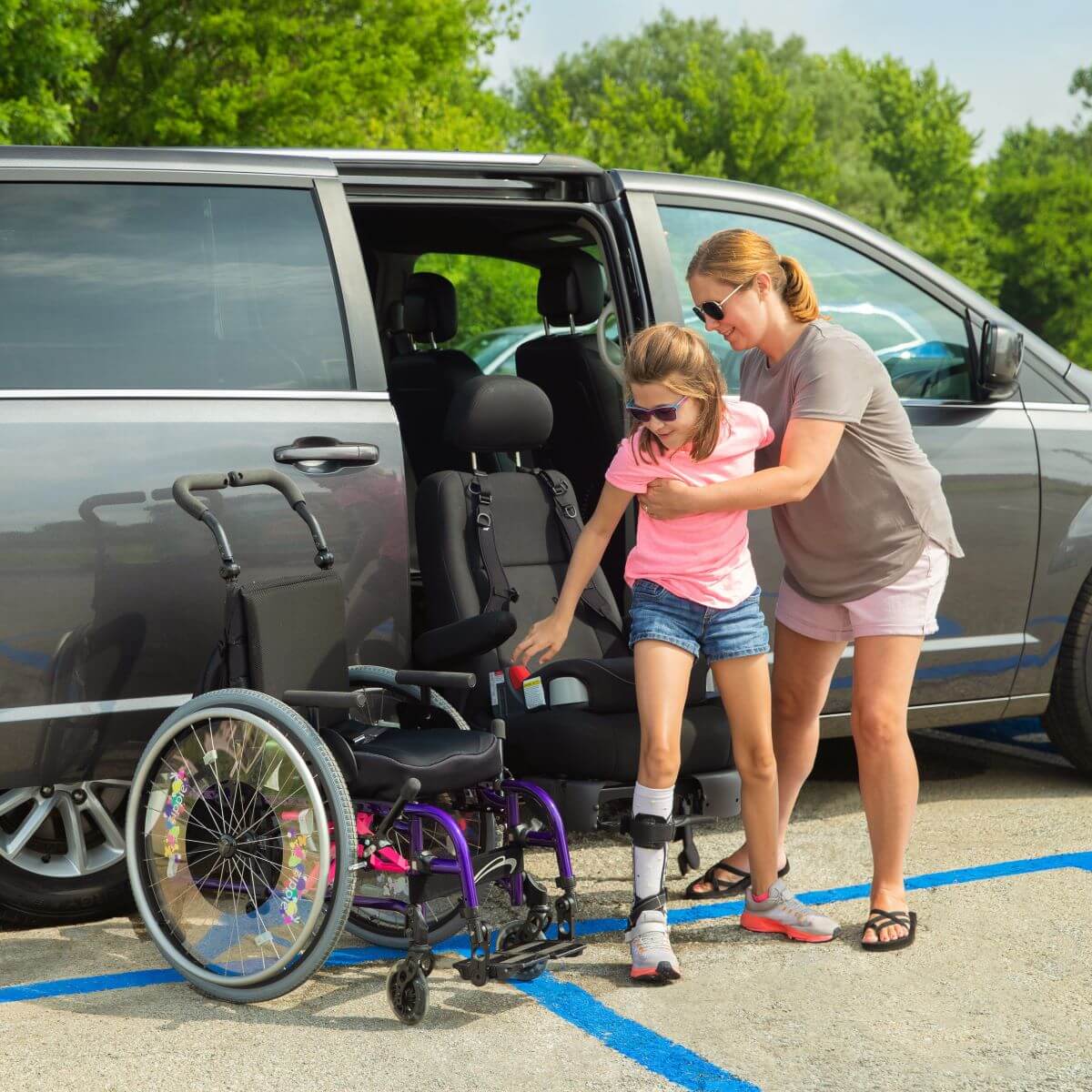 A woman assists a young girl with a leg brace as she stands up from her wheelchair beside a parked van in an accessible parking space.