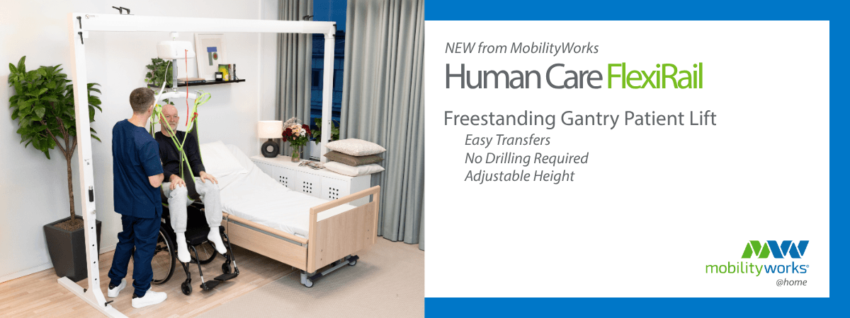 A caregiver uses a freestanding gantry lift to help a seated man transfer from a wheelchair to a bed in a well-lit bedroom.