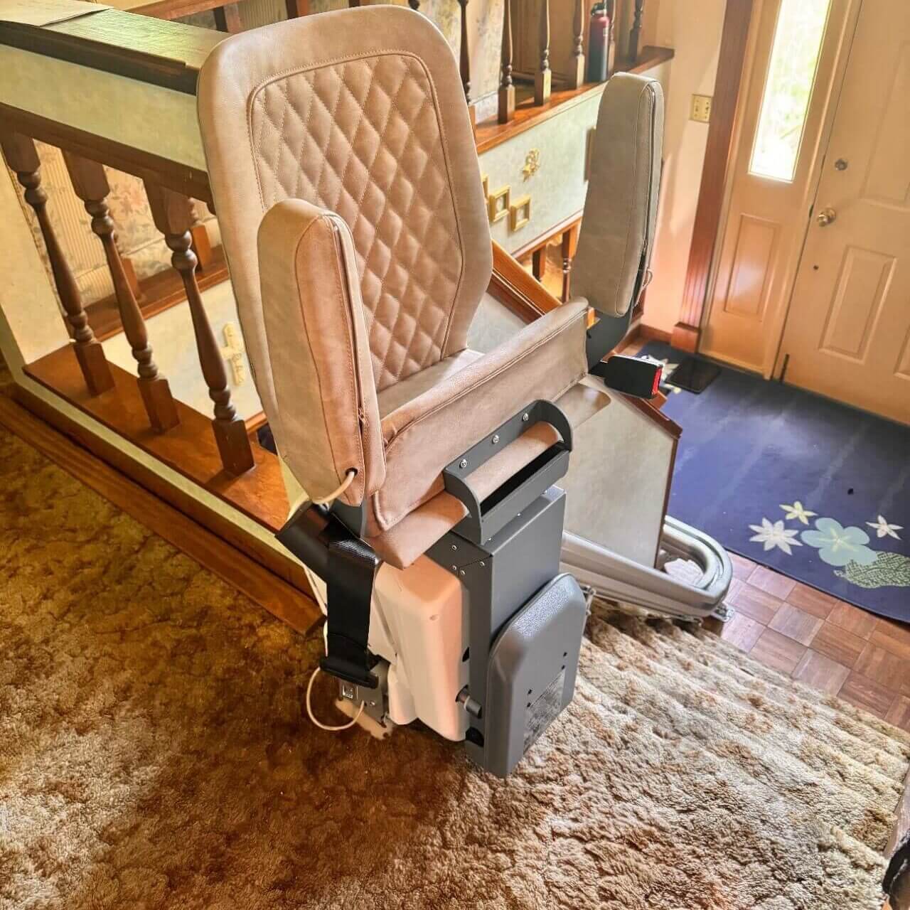 A stairlift installed on a carpeted staircase in a home, featuring a cushioned seat with armrests and tracks along the stairs.