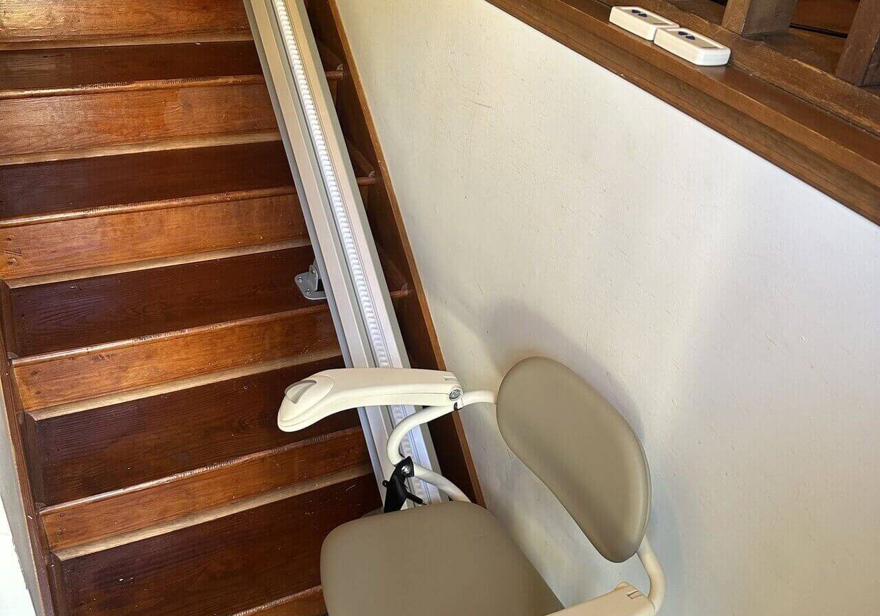 Stair lift chair installed on a wooden staircase with a beige seat and safety belt.