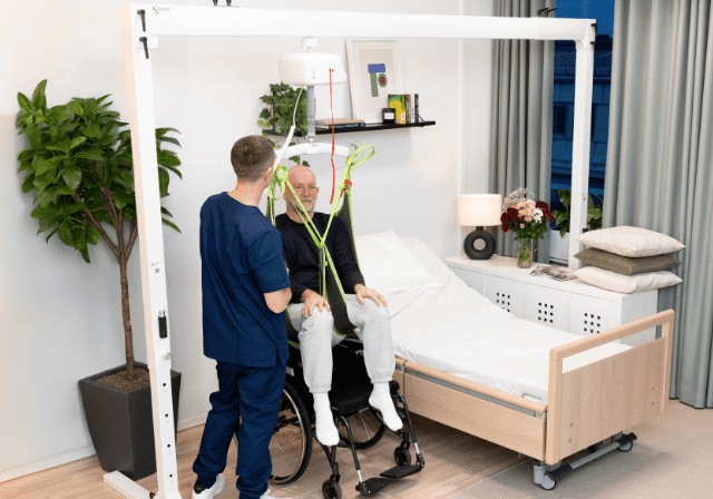 A nurse assists an elderly man in a wheelchair using a gantry patient lift next to a hospital bed in a modern, well-lit room.