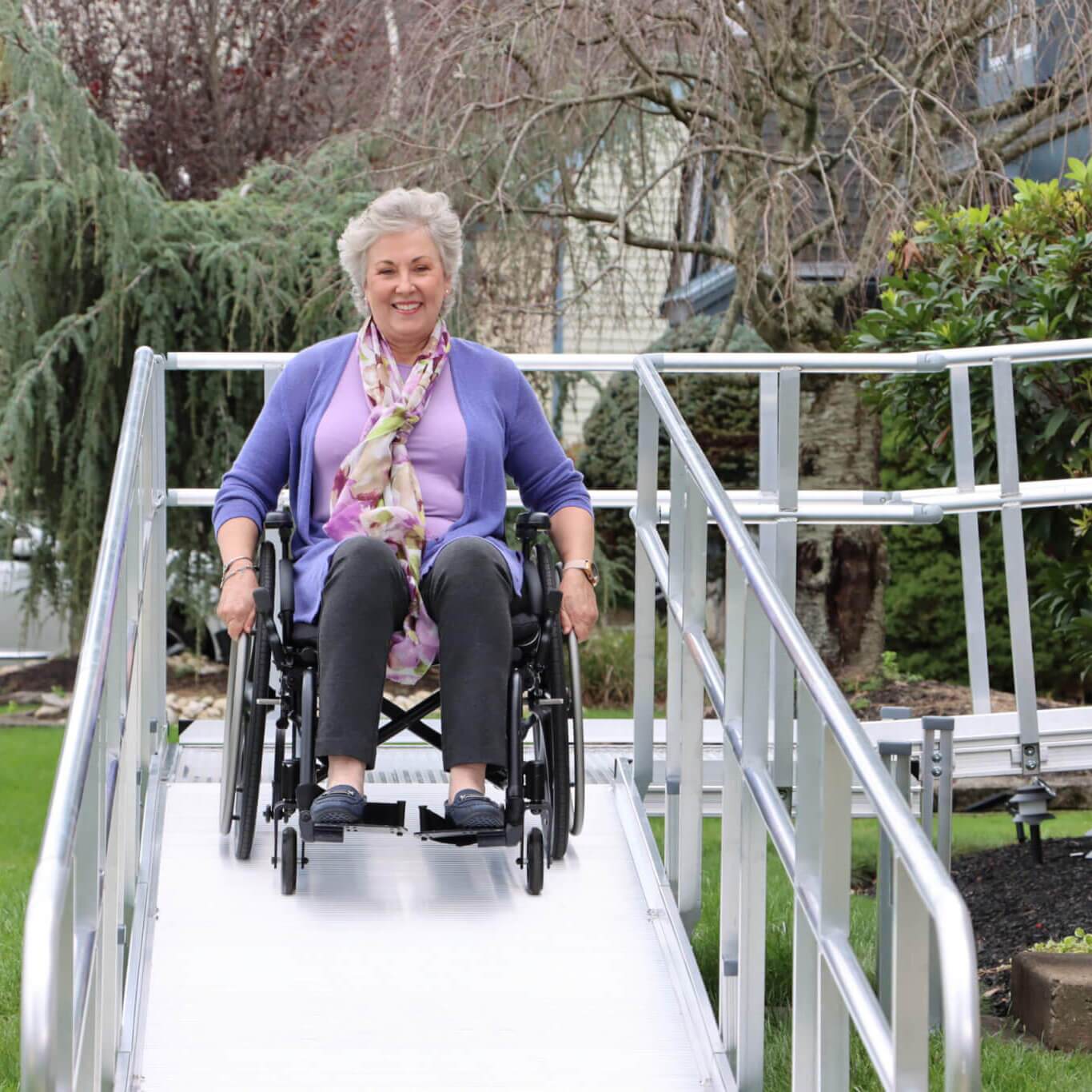 An older woman in a wheelchair moves down an outdoor metal accessibility ramp, smiling, with greenery and a silver car in the background.