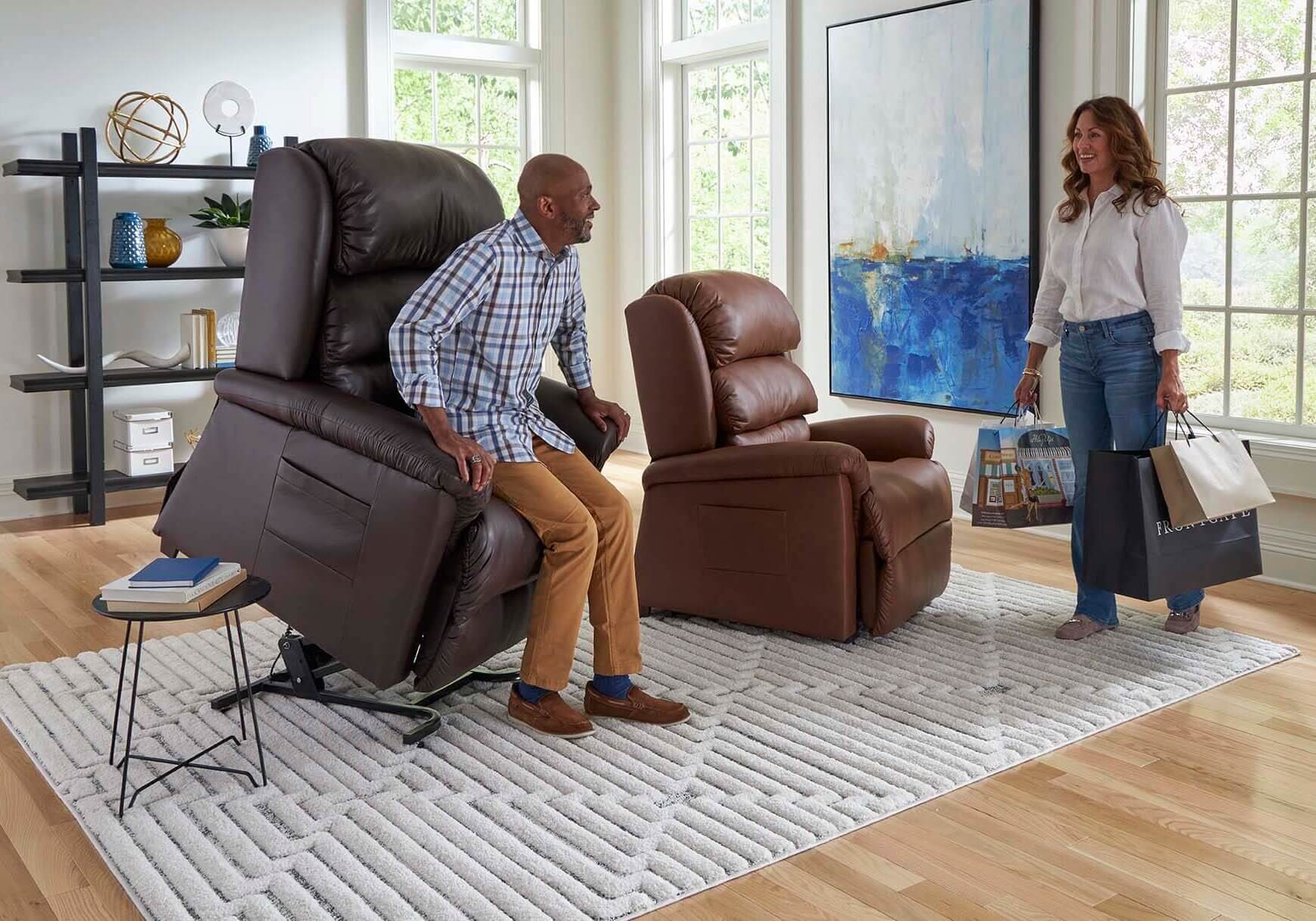 A man sits in a reclining chair in a living room, while a woman carrying shopping bags enters the room.