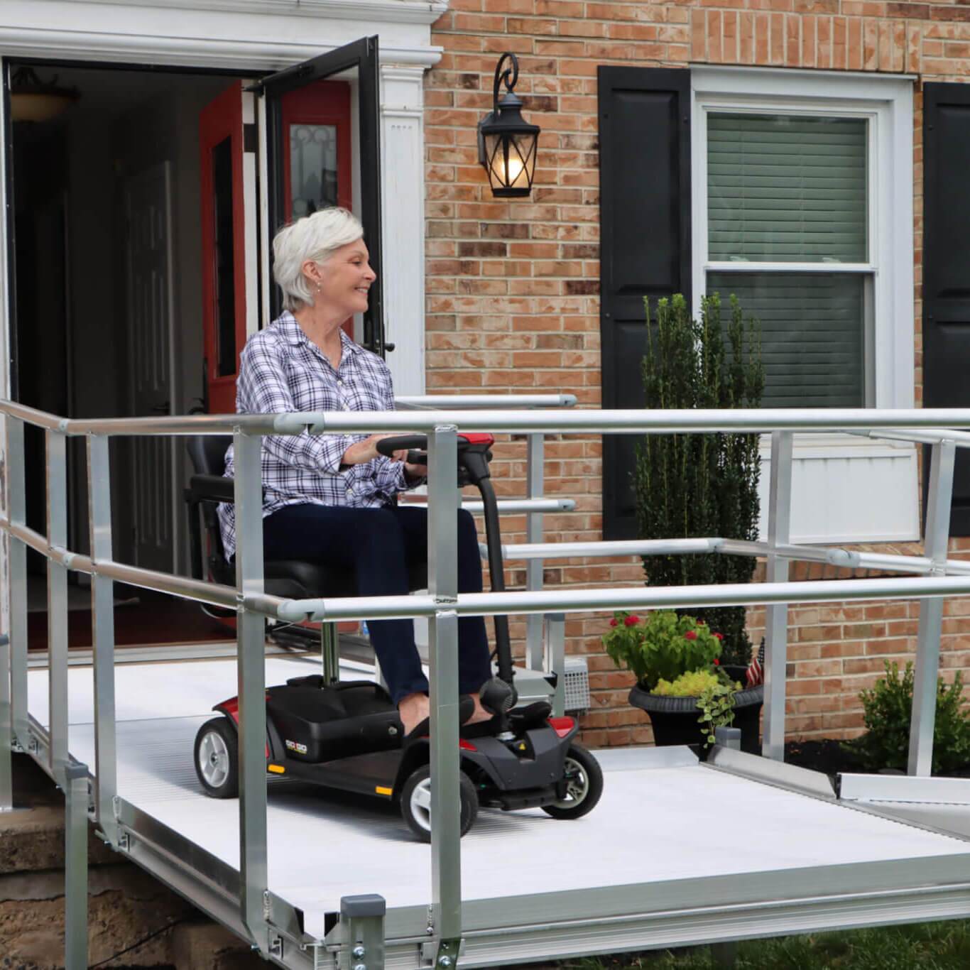 An older woman in a mobility scooter exits a brick house using an outdoor ramp with metal railings on a sunny day.