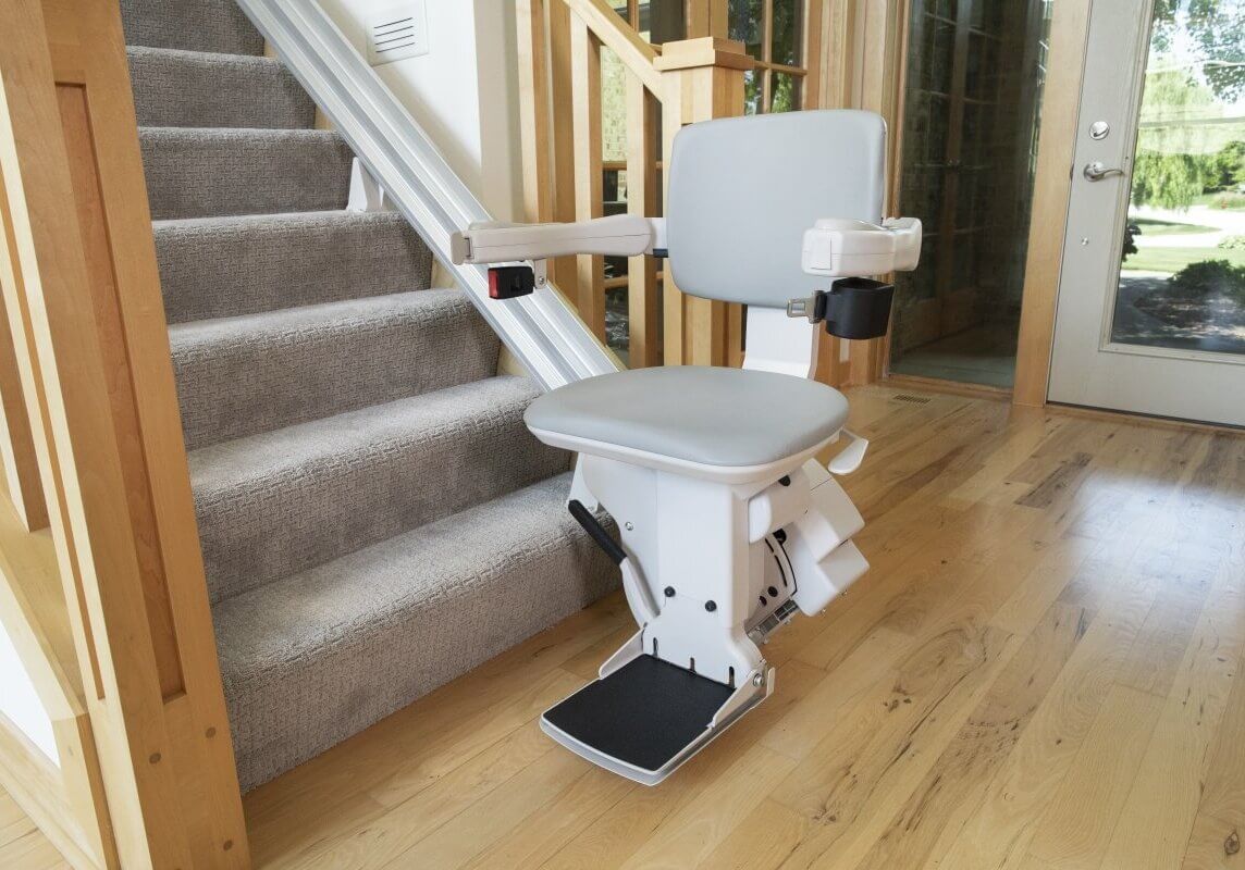 A stairlift is installed alongside a residential staircase, featuring a gray seat and armrests.