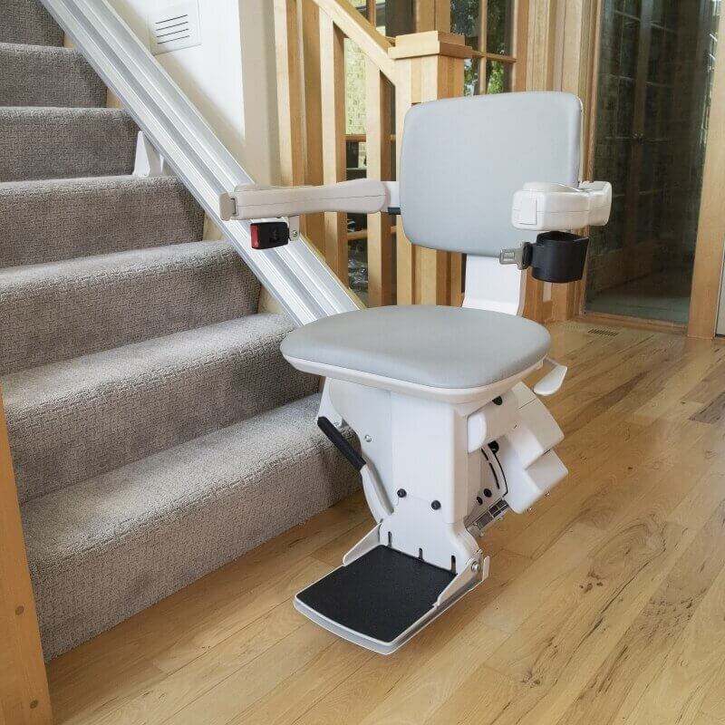 A stairlift is installed alongside a residential staircase, featuring a gray seat and armrests.