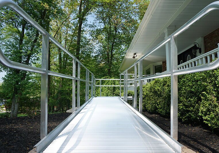 A metal accessibility ramp leads to the entrance of a house surrounded by greenery, seamlessly complementing the stairlifts inside.