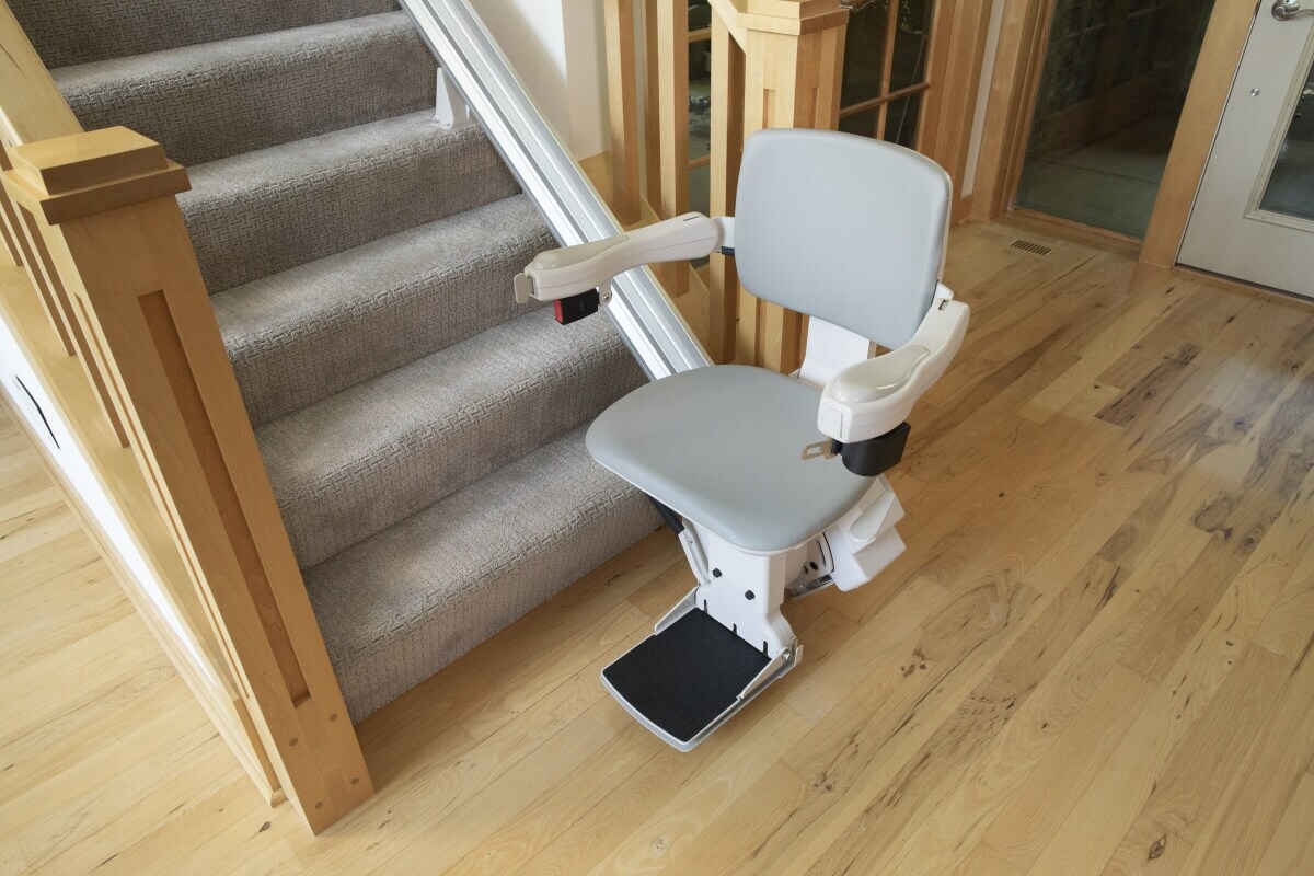 A stairlift with a gray seat is installed beside a carpeted staircase in a home with wooden floors and banisters.