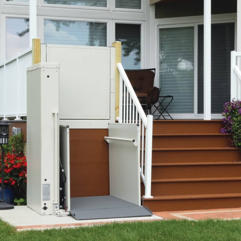 A residential porch lift is installed next to a set of stairs, leading up to a deck with a chair and potted plants nearby.