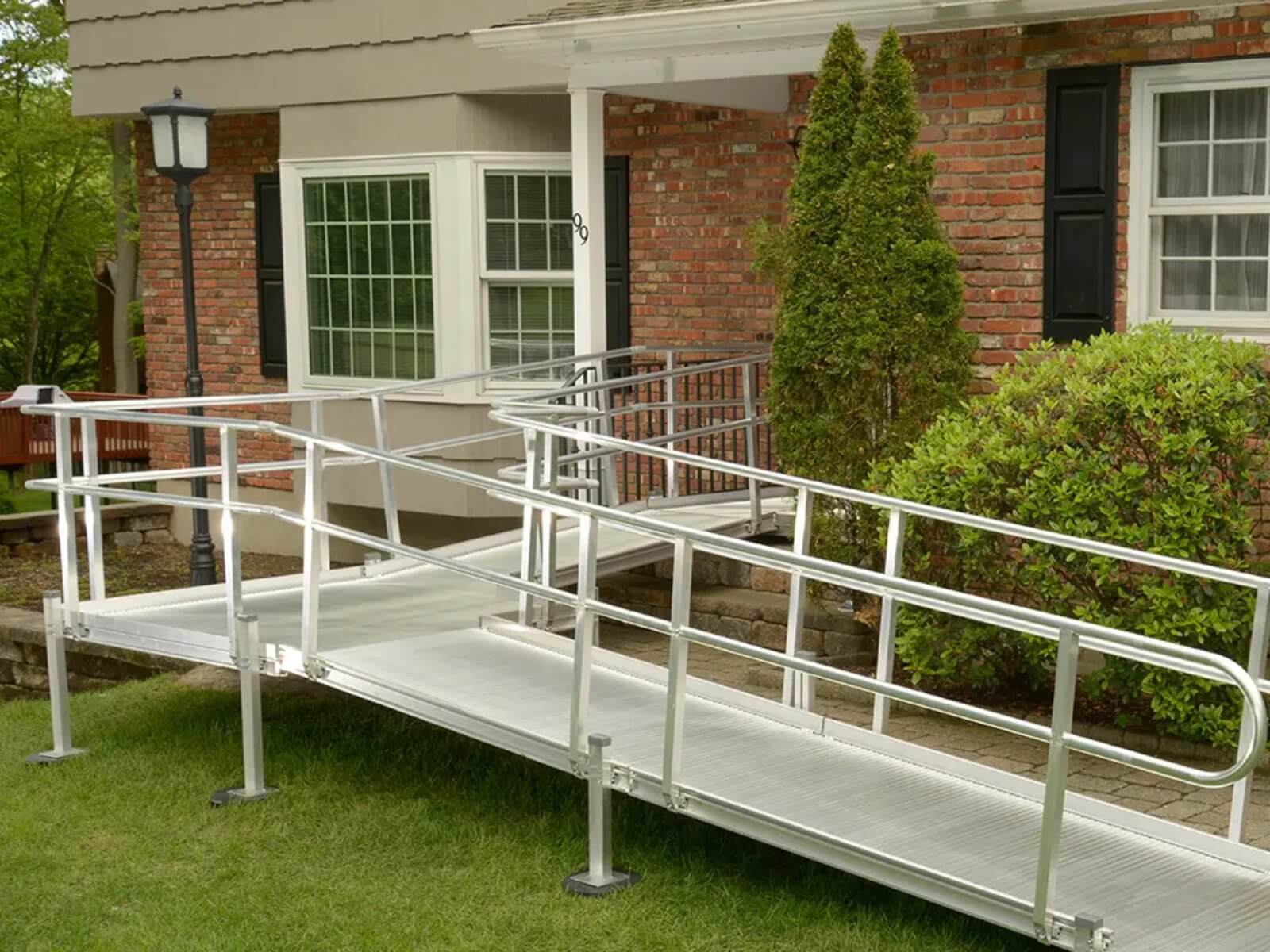 The aluminum wheelchair ramp for home leads gracefully to the entrance of a brick house, complemented by charming white windows and lush green shrubs.