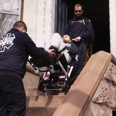 Two paramedics expertly maneuver a mobile stairlift, assisting a man on a stretcher as they navigate down a set of outdoor stairs.