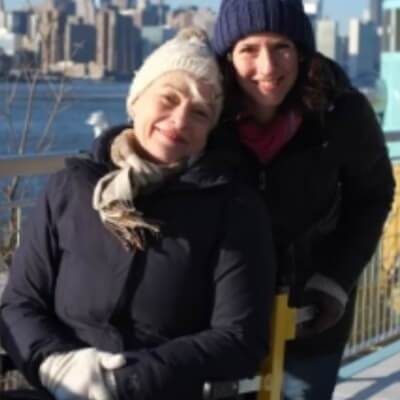 Two people in winter clothing smile outdoors, enjoying the crisp air. A city skyline and river provide a stunning backdrop, and nearby, a mobile stairlift offers accessibility for all to appreciate this view.