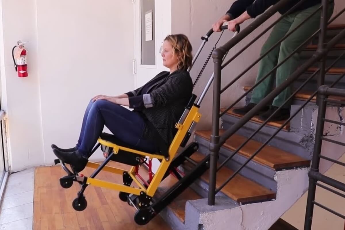 A person uses a yellow, mobile stair climbing wheelchair to descend stairs, assisted by another person controlling the chair from behind.