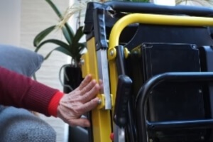 A person in a red sweater pushes a lever on a yellow, mechanical-looking mobile stairlift in a room with plants.