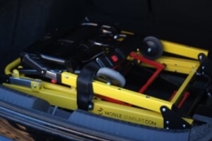 A folded yellow manual wheelchair, snugly secured with straps next to a compact mobile stairlift in the trunk of a vehicle.