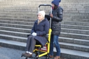 Two people are outdoors; one is seated in a mobile stairlift wheelchair, while the other stands behind, expertly operating the device near a flight of steps.